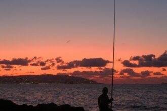 Man Sits with Fishing Road by Ocean at Sunset